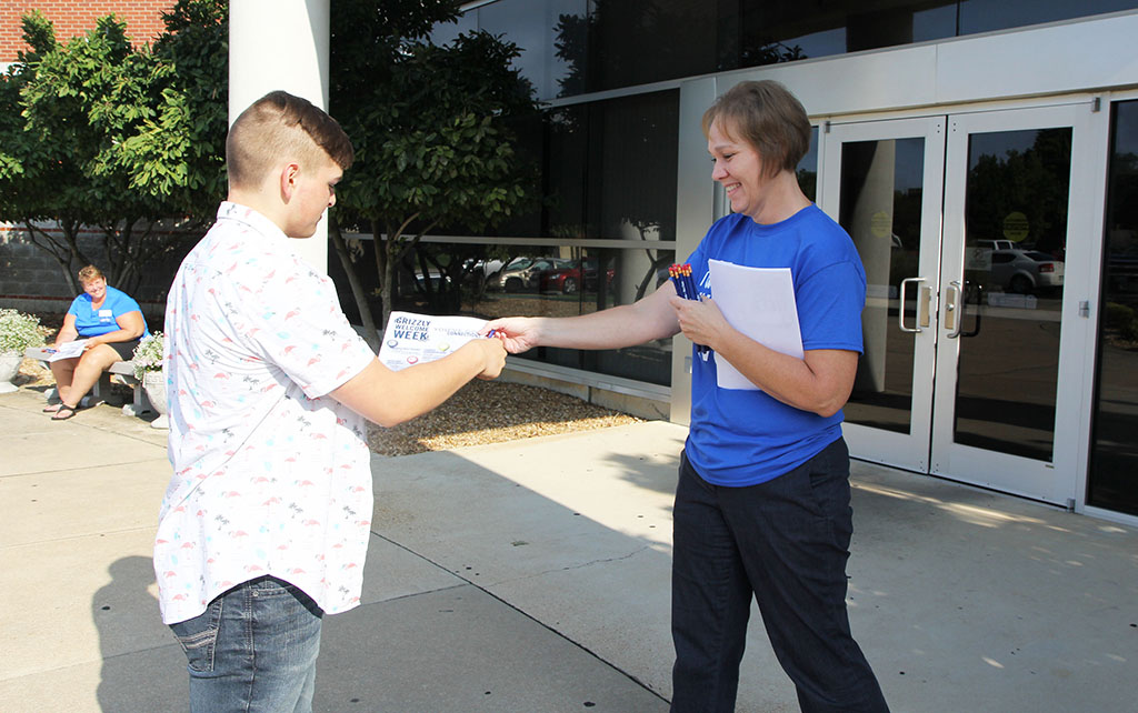 Two people are standing on a sidewalk in front of the doorway to a building. A woman hands a flyer and a pencil to a young man, welcoming him to the campus. Dr. Angela Totty, dean of student services, welcomes freshman Robert Cavitt, West Plains, to his first day of classes Monday at Missouri State University-West Plains. Several faculty and staff members manned doorways to the campus’ main classroom buildings on the first day to welcome students and give them information about upcoming events as part of Operation Smile. (Missouri State-West Plains Photo)