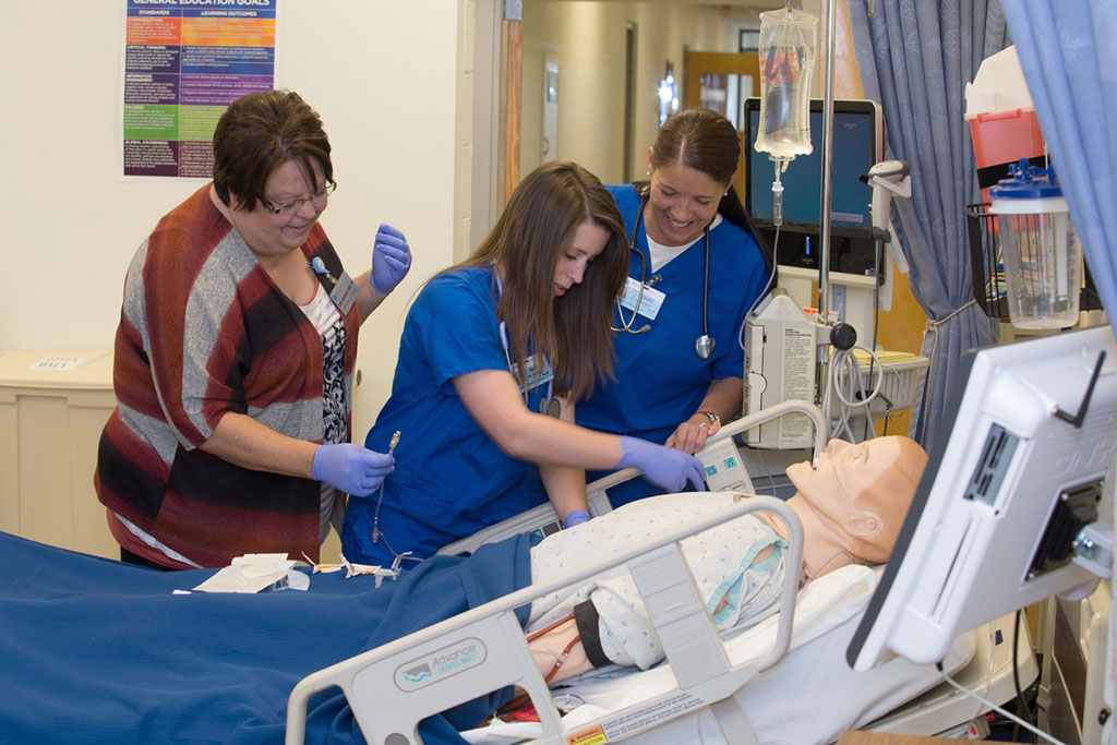 Nursing students insert an IV in a training dummy in a simulation lab with guidance from an instructor.