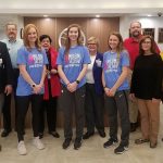 A group of people representing Ozarks Medical Center, Community First Banking Company and Grizzly Volleyball stand in a row in the OMC lobby. The volleyball representatives are wearing the T-shirts being sold for the fundraiser.