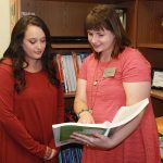 The student and instructor look through a textbook while standing in the instructor's office.