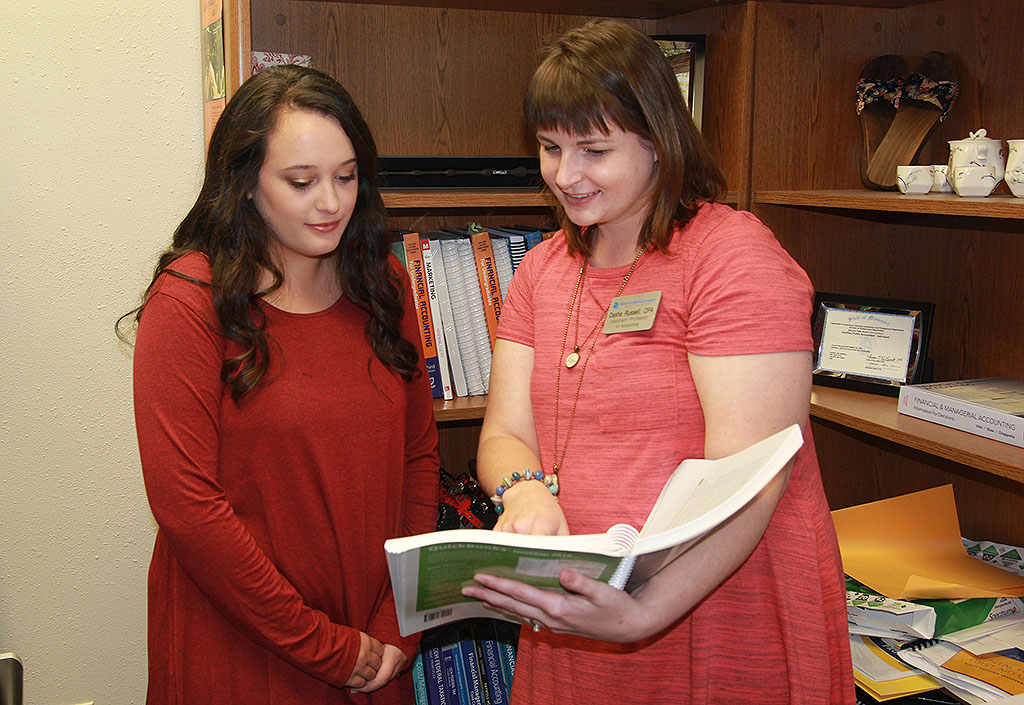 The student and instructor look through a textbook while standing in the instructor's office.