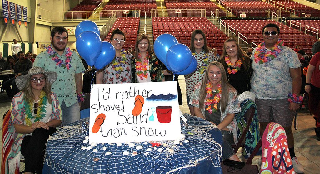 Team members wearing colorful Hawaiian shirts are sitting and standing around a table decorated things commonly found on a beach and with balloons. 