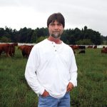 Teddy Gentry stands in the middle of a field with cattle behind him.