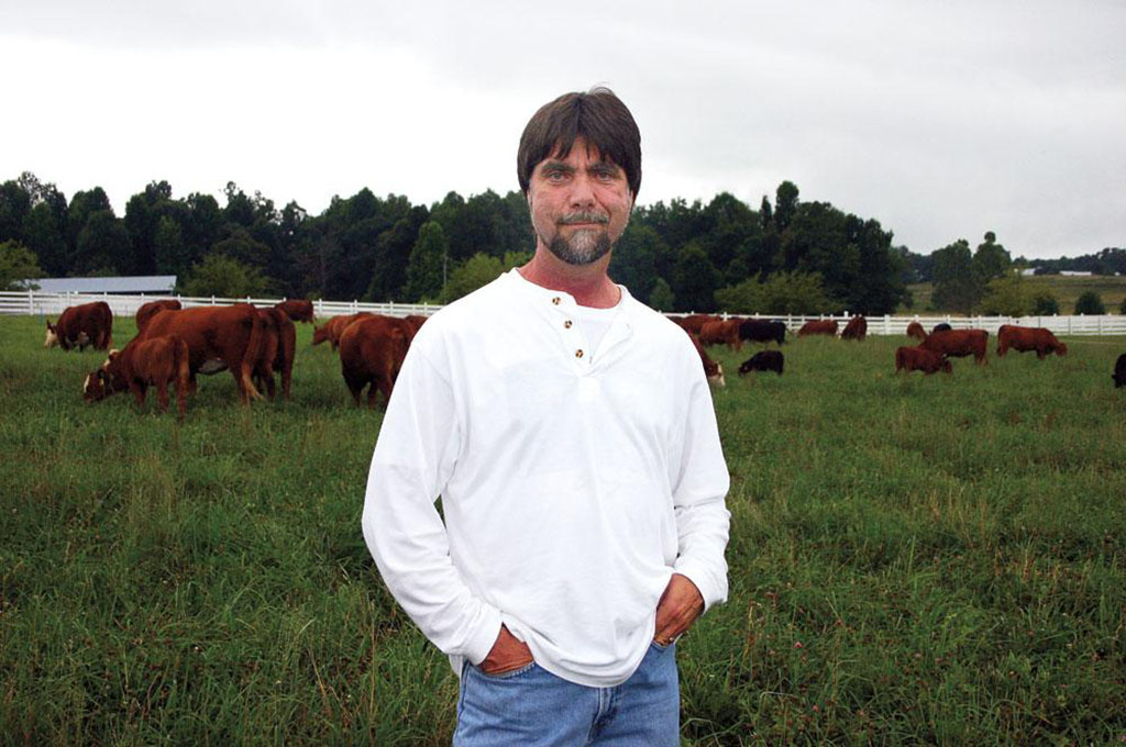 Teddy Gentry stands in the middle of a field with cattle behind him.