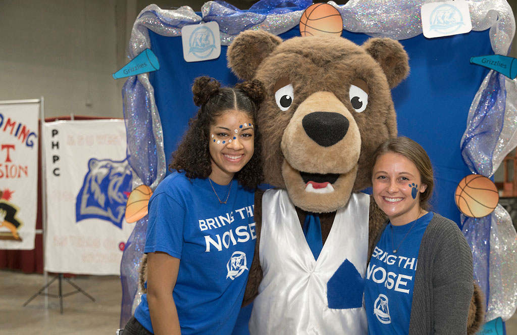 Two students with faces painted for homecoming stand with the Grizzly mascot in front of a blue backdrop.