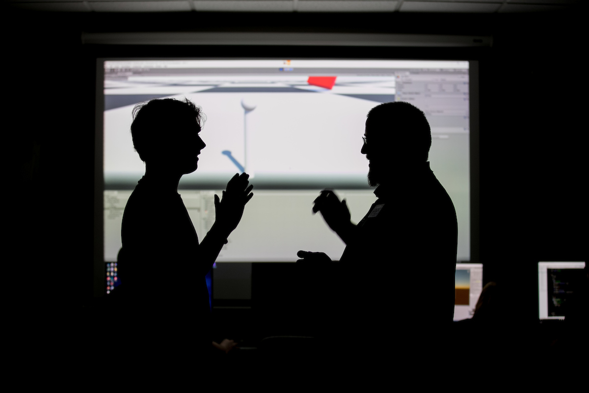 A student and teacher stand silhouetted in front of a screen showing a scene from a video game.