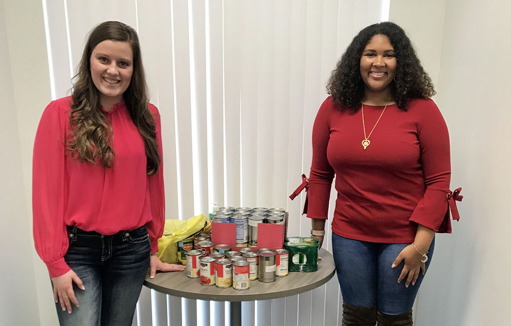 The two ladies stand next to a table with several canned good items sitting on it.