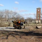 A man driving a small bulldozer and another man set posts for a fence. In the background is the campus' bell tower.