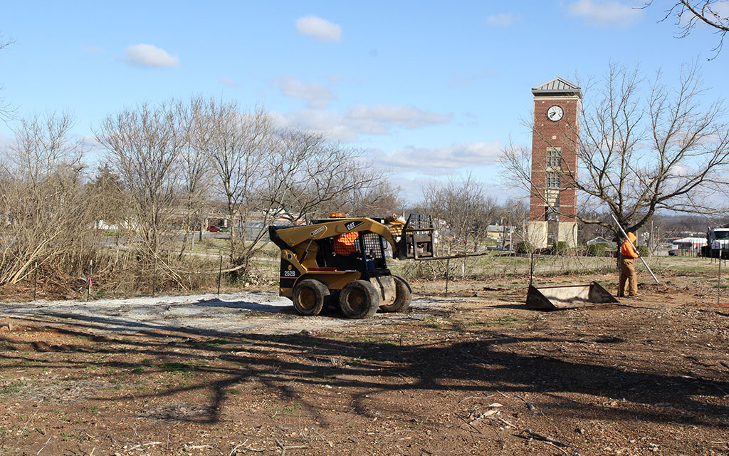 A man driving a small bulldozer and another man set posts for a fence. In the background is the campus' bell tower.