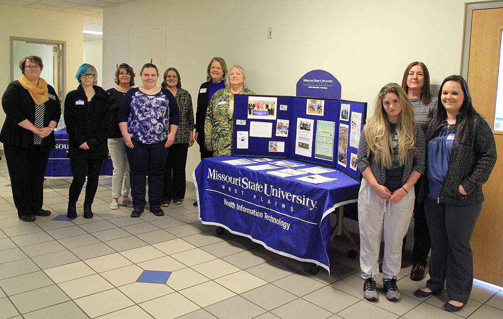 The group stands in rows next to a table with information about Health Information Technology.