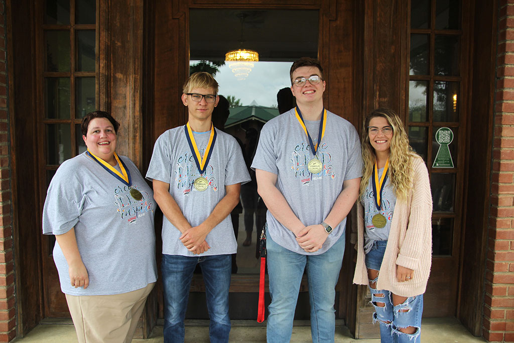 Four students stand in a row in front of a glass door entrance to a building.