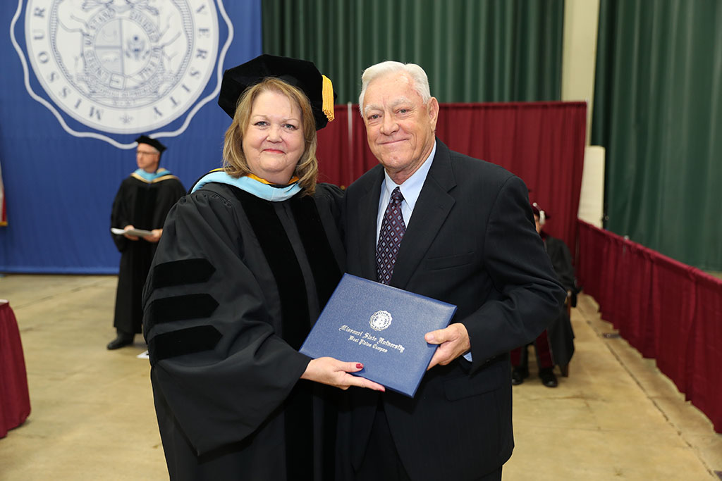 Two people stand next to each other holding an engraved diploma folder.