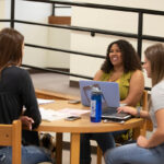 Three students are sitting around a table talking and studying with their books and laptops.