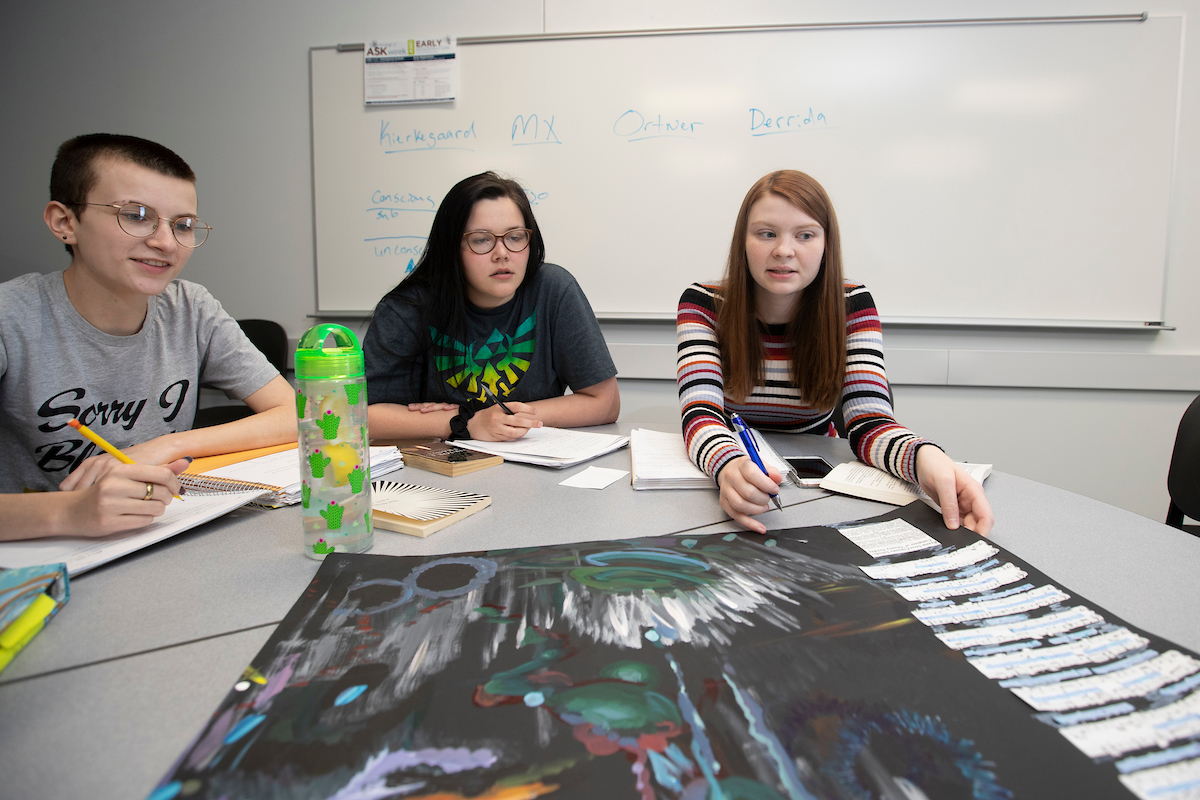 Students seated around a table in a classroom discuss a poster that is part of a class project.