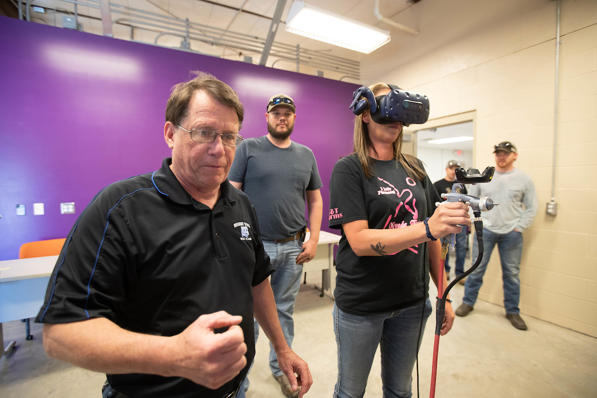 An instructor shows a student wearing goggles and holding a spray paint gun how to use the virtual reality computer program.