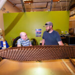 Teacher and student stand behind a piece of equipment and hold up a large lattice plank created on the machine.