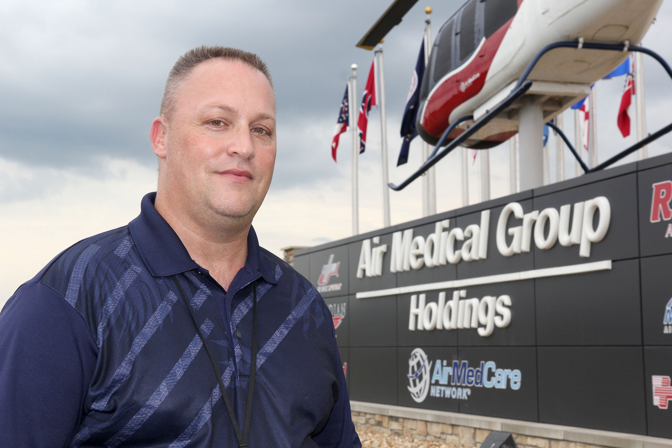 A man stands outside in front of a sign that says Air Medical Group Holdings and Air Med Care. On top of the sign are flags from different states and a medical helicopter.