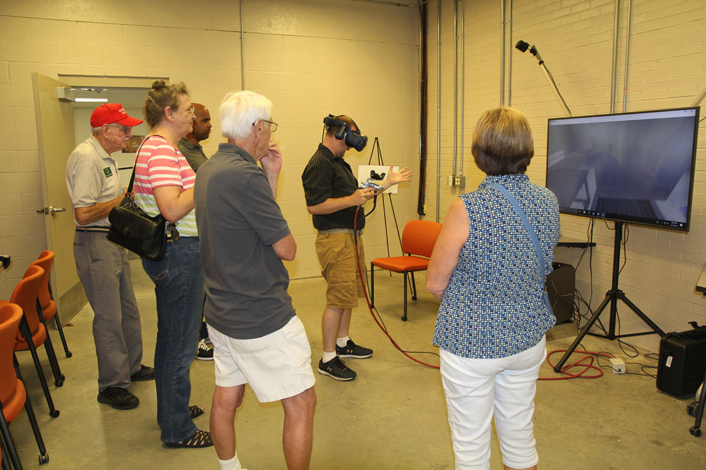 A group of people are gathered around a man wearing virtual reality goggles while pointing a paint gun at a big screen TV.