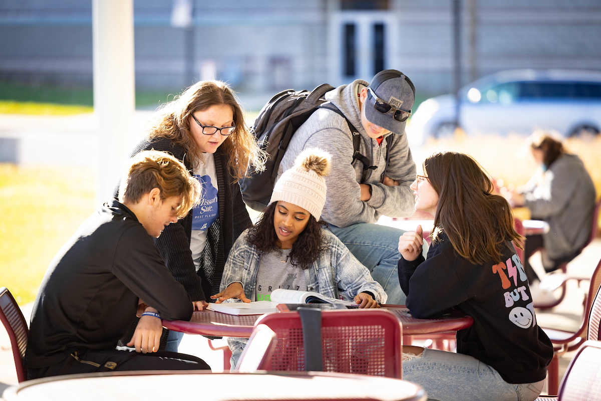 Students gathered around an outdoor table looking at a textbook