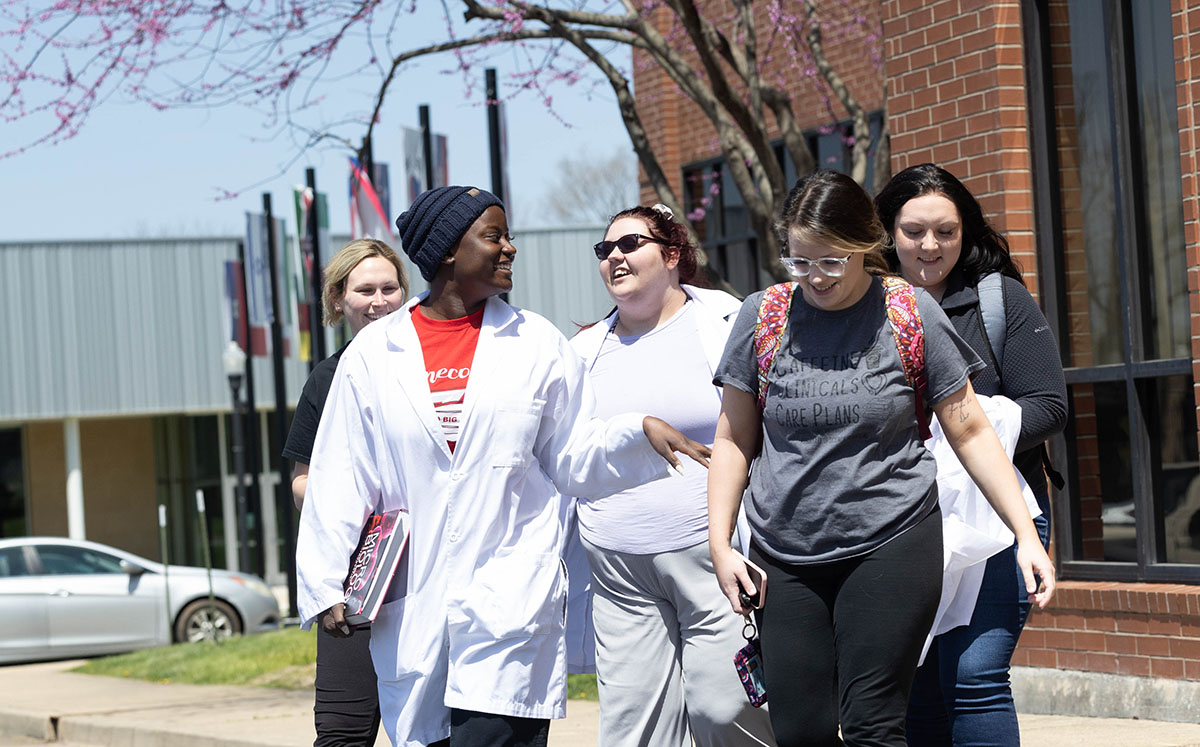 Five female students with backpacks, books and some wearing lab coats are talking as they walk across campus.