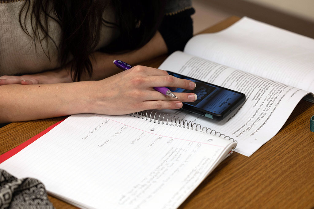 A notebook and textbook are open on a desk. A student is using a device sitting on top of the textbook.