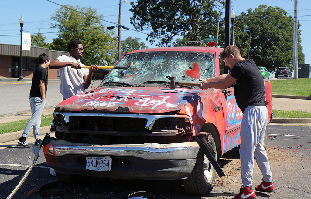 Two young men stand on either side of an old pickup truck. Each is hitting the truck with a sledge hammer.
