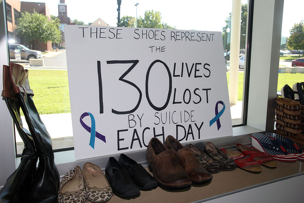 Pairs of shoes sit on a window sill. Behind them a handmade poster reads "These shoes represent the 130 lives lost by suicide each day."