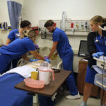 Four nursing students gathered around a hospital bed practicing resuscitation procedures on a medical mannequin.