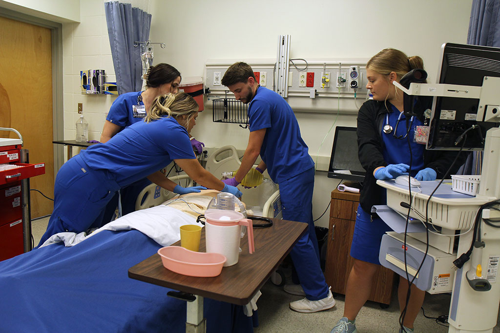 Four nursing students gathered around a hospital bed practicing resuscitation procedures on a medical mannequin.