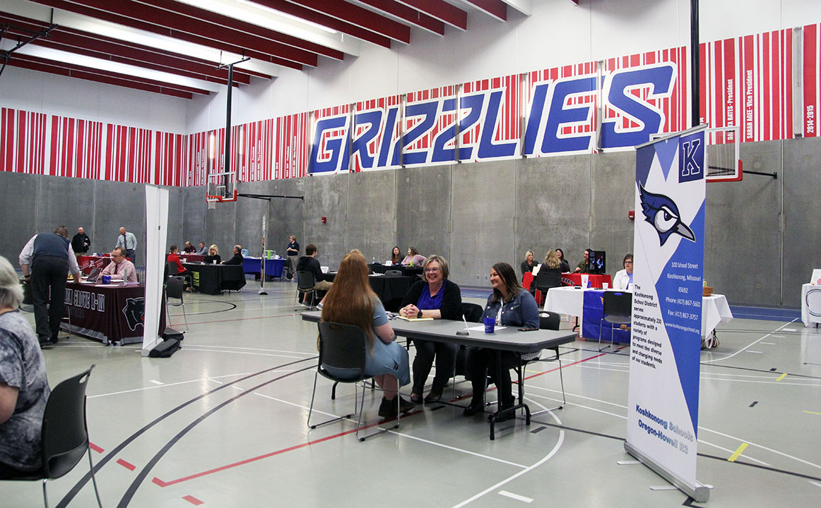 A large room is filled with tables. People are sitting or standing at each table and talking.