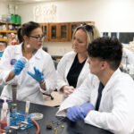 An instructor discusses a biology experiment with two students at a lab table. All three are wearing white lab coats. The instructor, standing left, is holding a petri dish.