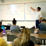 A teacher stands at a white board showing mathematical problems to a classroom