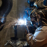 A person in welding gear, including gloves and mask, welds a piece of metal while sparks fly from the weld.