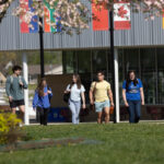 A group of five students walk across the lawn talking to each other. In the background is a building and flag poles with colorful flags attached.