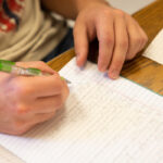 A student writes math equations on a lined piece of notebook paper. Only the student's hands are shown.