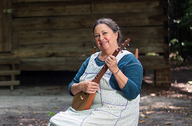 A woman in old-time clothes sits on a wooden chair holding a stringed instrument in front of an old log cabin.