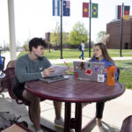 Two students sit at a table outside with their laptops and notebooks. In the background, a student walks on a sidewalk underneath flag poles.